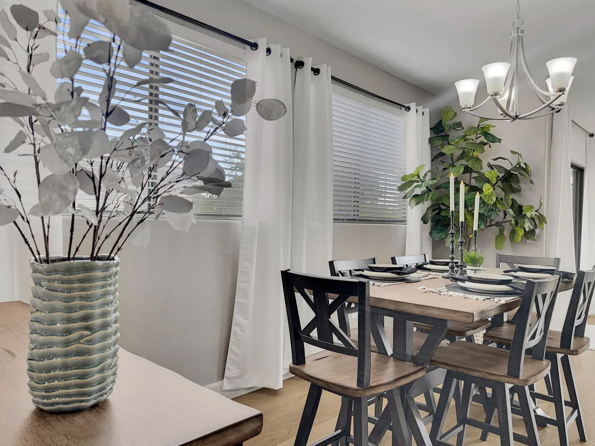 A warm, inviting dining area featuring a large table with multiple chairs, a decorative vase centerpiece, hanging chandelier, and lush green plant. The setting is ideal for shared meals, celebrations, or family board game nights, capturing the essence of togetherness.