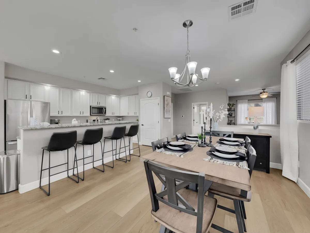 A warm, open-plan dining area set for a meal, with a glimpse of the kitchen countertop and a sparkling chandelier overhead. The scene radiates comfort and ease, ideal for gatherings and everyday living.