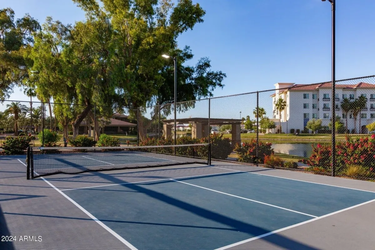 A vibrant exterior shot showing a tennis court framed by lush trees and a nearby building, with people engaged in active play and others relaxing or socializing in the shaded areas. The scene is energetic yet inviting, highlighting additional leisure amenities within the community.