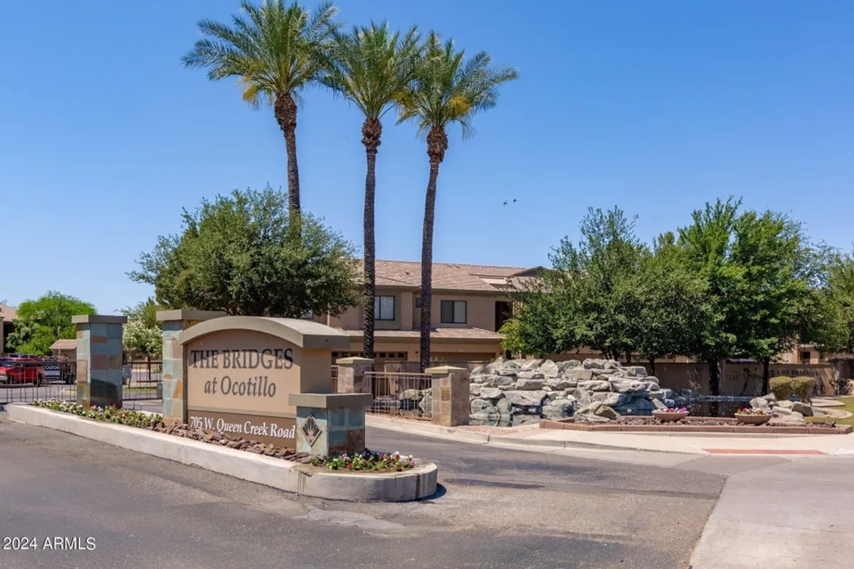 An inviting exterior image showcasing an entrance sign flanked by palm trees and landscape rocks, with a charming water feature in the background. People are seen strolling or relaxing by the water, creating a social yet tranquil atmosphere that emphasizes community and the connection to nature.