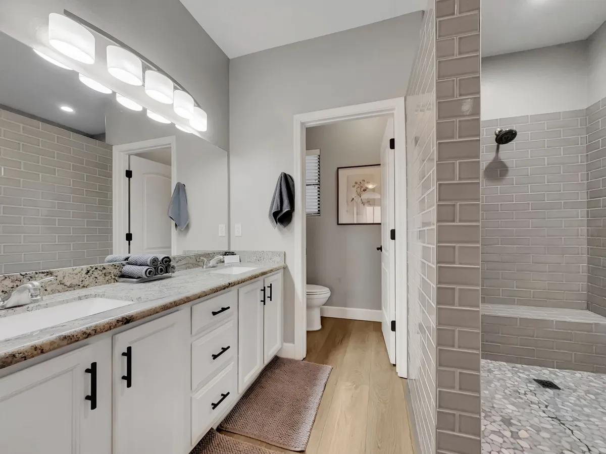 A modern bathroom with a double vanity, granite countertop, large mirror, and stylish decorative elements. The shower area is visible, highlighting both function and luxury for guests.