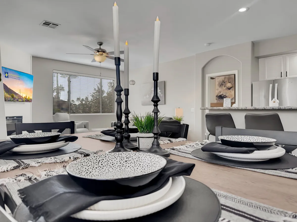 A bright dining area set up for a meal, featuring a dining table with decorative plates, black candlesticks, a wall-mounted TV, and ample natural light streaming through a window. The arrangement is both intimate and welcoming, ready for gathering or special celebrations.