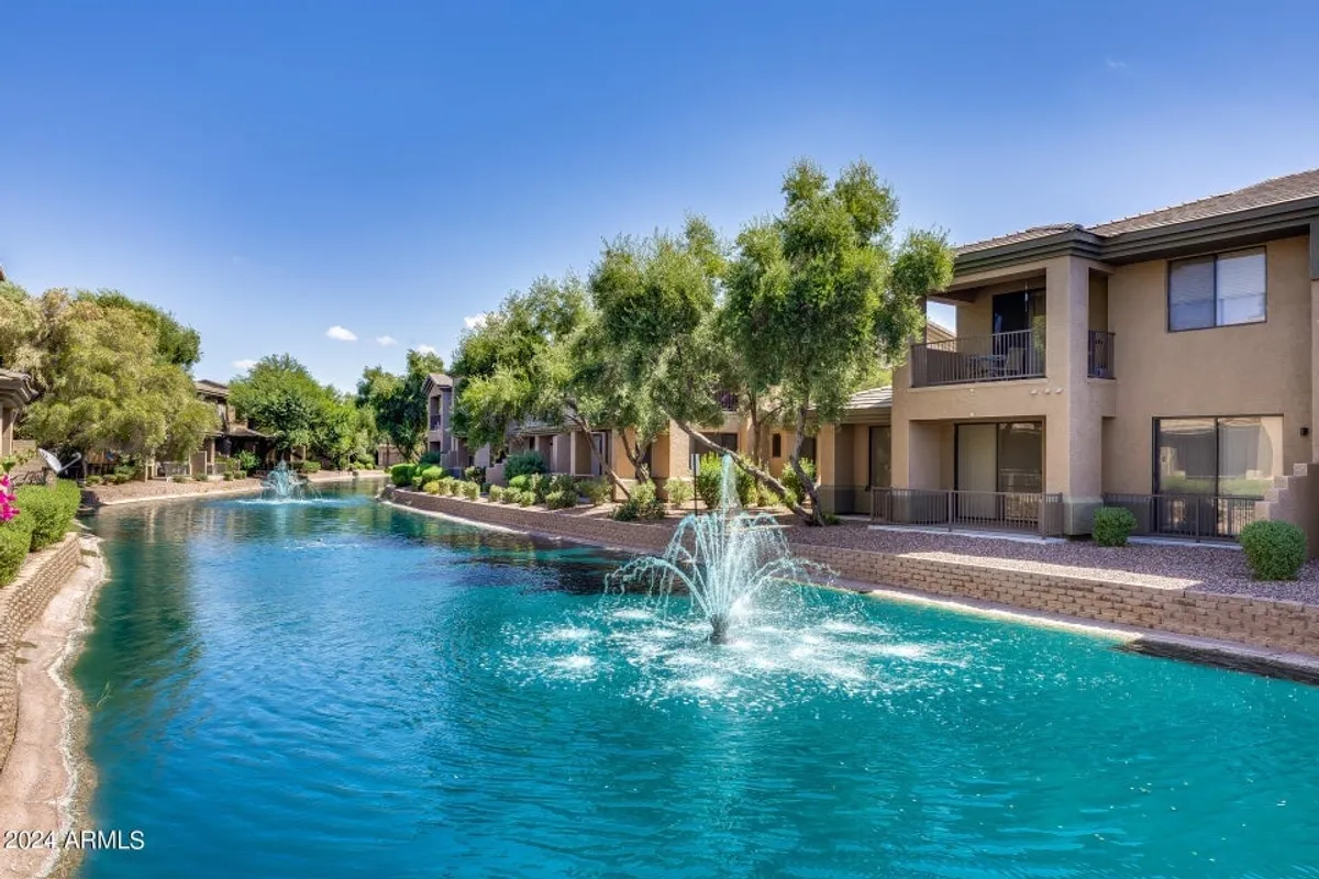 A tranquil exterior scene featuring a sparkling water fountain surrounded by lush greenery and well-maintained walking paths. Elegant apartment buildings border the area, with balconies overlooking the serene waterway. The paths invite leisurely strolls, while the water reflects the warm sunlight, enhancing the atmosphere of relaxation and natural beauty.
