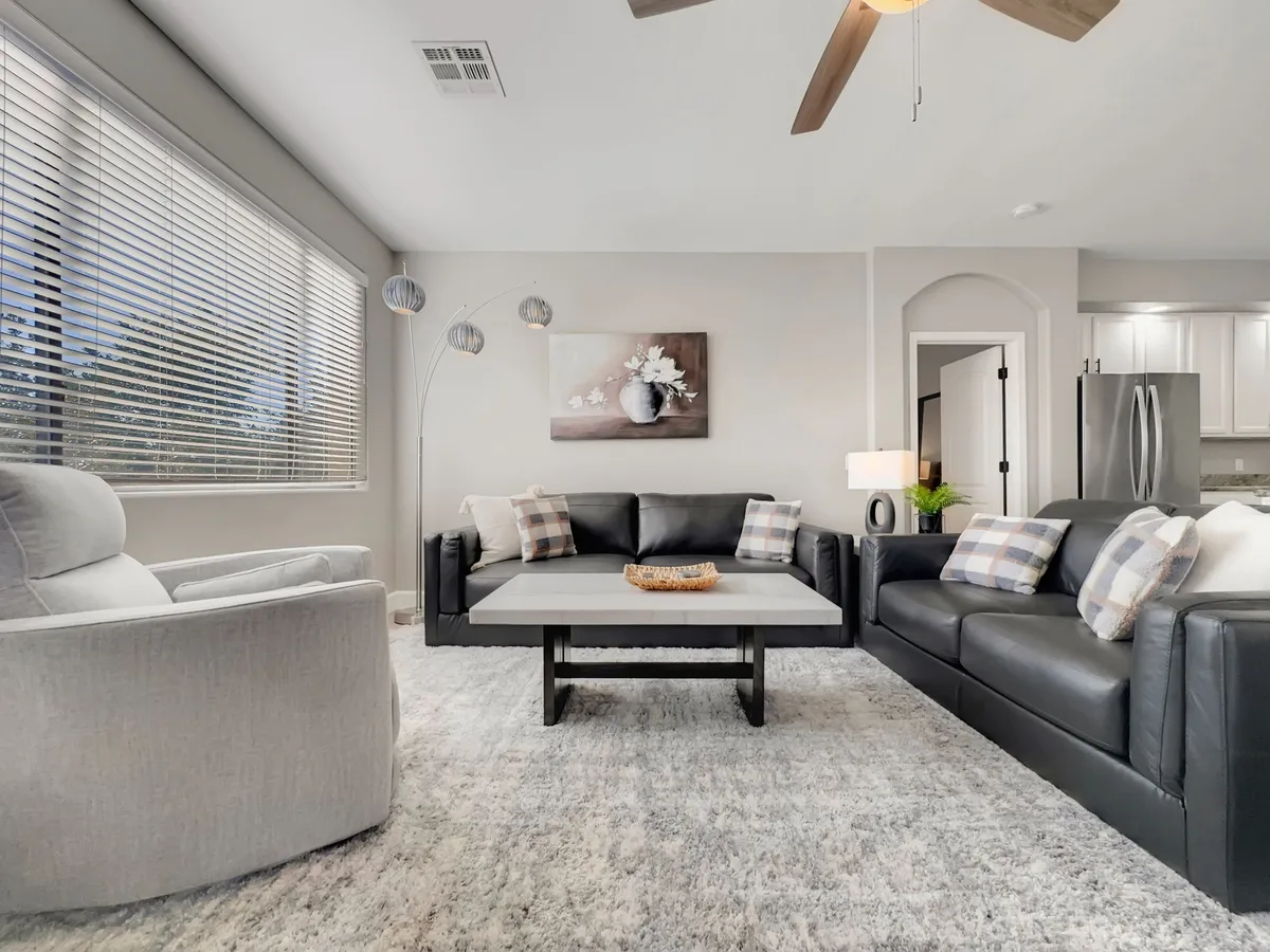 A cozy living room scene features a comfortable sofa, a sleek coffee table, tasteful artwork, and a modern floor lamp. The window in the background hints at daylight outside, providing a sense of connection to the world beyond the condo. The room is arranged to invite relaxation and socializing after a busy day.