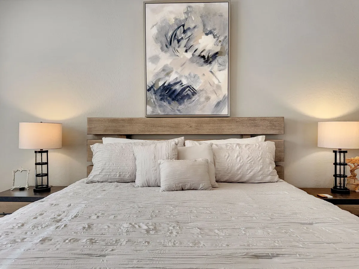 This image features a serene bedroom with white bedding, an abstract painting above the bed, and a nightstand with lamps. Soft natural light pours in, creating a tranquil atmosphere ideal for restful escapes.